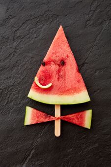 A Juicy Piece Of Watermelon On A Stick In The Form Of A Smiling Face On A Dark Slate Background With Copy Space. Flat Royalty Free Stock Photo A Juicy Piece Of Watermelon On A Stick In The Form Of A Smiling Face On A Dark Slate Background With Copy Space. Flat Royalty Free Stock Photo