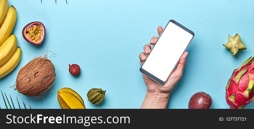 A variety of tropical fruits and a female hand with a phone on a blue background with copy space. The concept of buying products on the Internet. Flat lay. A variety of tropical fruits and a female hand with a phone on a blue background with copy space. The concept of buying products on the Internet. Flat lay