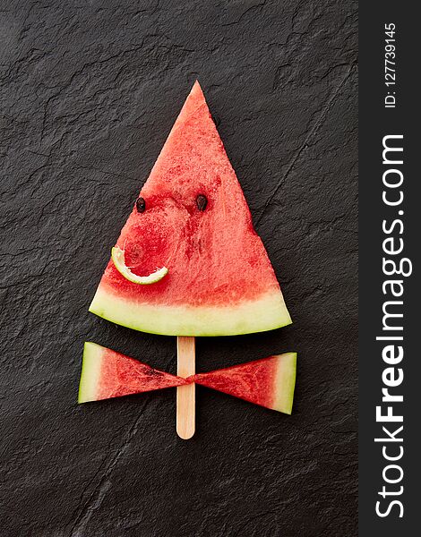 A juicy piece of watermelon on a stick in the form of a smiling face on a dark slate background with copy space. Flat
