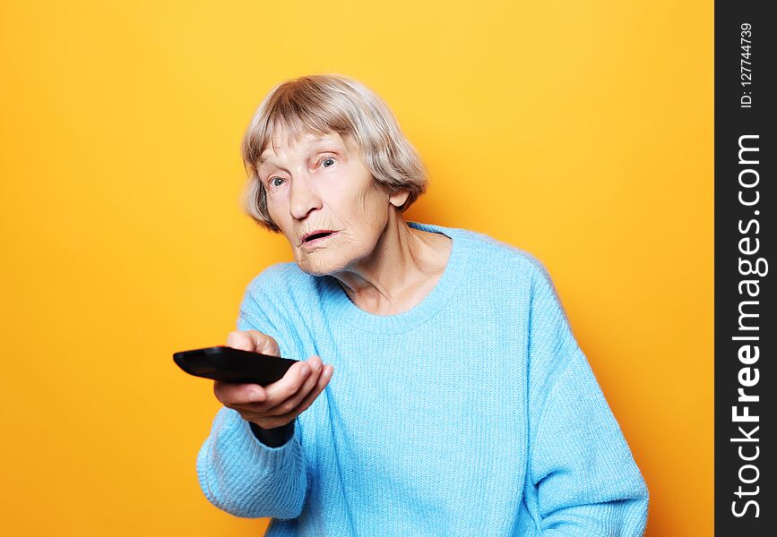 Lifestyle and people concept: funny grandma is holding a TV remote over yellow background