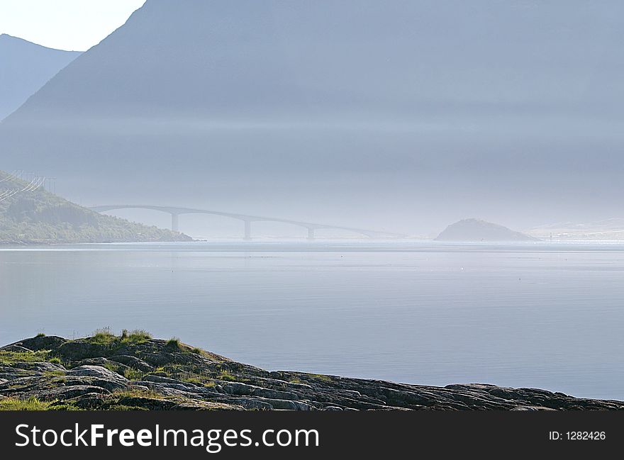 View from the south coast of the island Vestvagøya on the Vestfjords and the bridge between this island and Austvagøya; both islands are part of the Lofoten in Norway. This photograph has been made in the early morning if the mist is solved by the sun. View from the south coast of the island Vestvagøya on the Vestfjords and the bridge between this island and Austvagøya; both islands are part of the Lofoten in Norway. This photograph has been made in the early morning if the mist is solved by the sun.