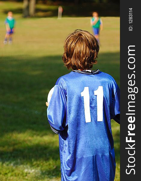 Young boy playing soccer on s sunny afternoon. His back is to the camera. There are two other players visible in the blurred background. Young boy playing soccer on s sunny afternoon. His back is to the camera. There are two other players visible in the blurred background.
