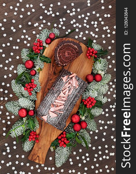 Chocolate log yule cake with winter flora of snow covered fir, holly and mistletoe on rustic wooden board and oak table background with snowflake sprinkles. Top view. Chocolate log yule cake with winter flora of snow covered fir, holly and mistletoe on rustic wooden board and oak table background with snowflake sprinkles. Top view.