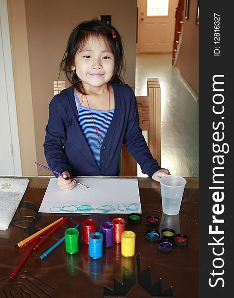 Asian girl painting at a table with paints in front of her and a paint brush inside her hand. Asian girl painting at a table with paints in front of her and a paint brush inside her hand