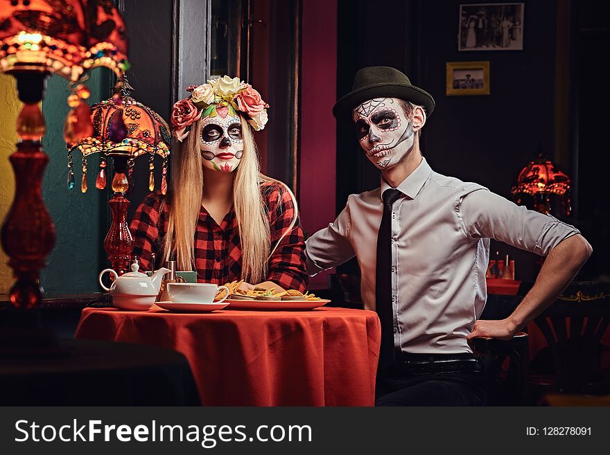 Halloween and Muertos concept. Young attractive couple with undead makeup ordered nachos during dating at a mexican restaurant. Halloween and Muertos concept. Young attractive couple with undead makeup ordered nachos during dating at a mexican restaurant.