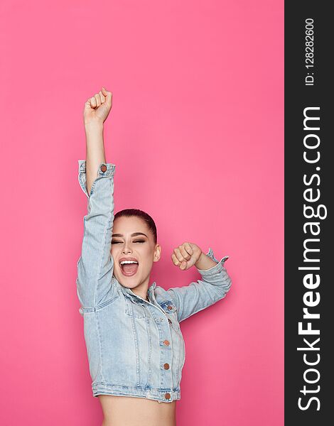 young woman in jeans jacket is holding arm raised and shouting. Waist up studio shot on pink background. young woman in jeans jacket is holding arm raised and shouting. Waist up studio shot on pink background.