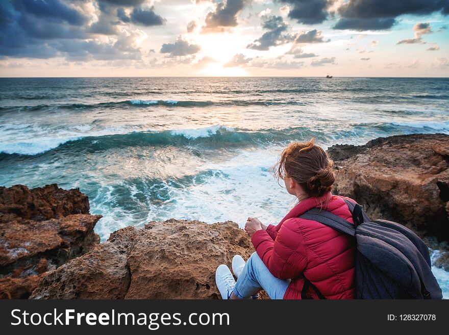 Young woman traveler sits on the shore of the winter sea and watches the bright colorful sunset