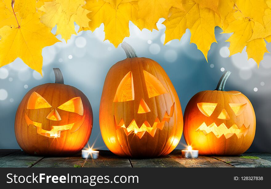 Three Halloween pumpkins head jack o lantern and candles on wooden table background , bokeh lights and autumn yellow maple leaves frame. Three Halloween pumpkins head jack o lantern and candles on wooden table background , bokeh lights and autumn yellow maple leaves frame