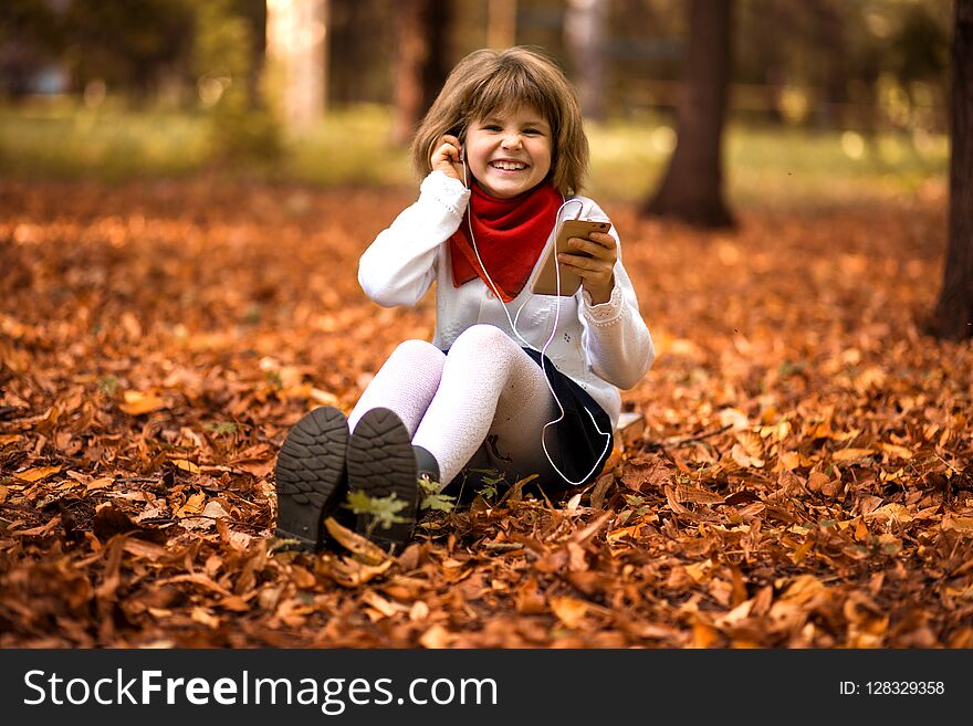 Happy little girl sits on yellow leaves and listens to music in autumn