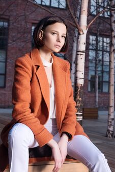 Young Woman In Red Coat And White Pants Sitting On A Wooden Bench. Street Royalty Free Stock Images