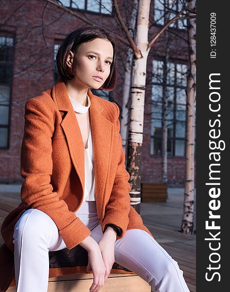 Young woman in red coat and white pants sitting on a wooden bench. Street fashion closeup portrait. Business urban style