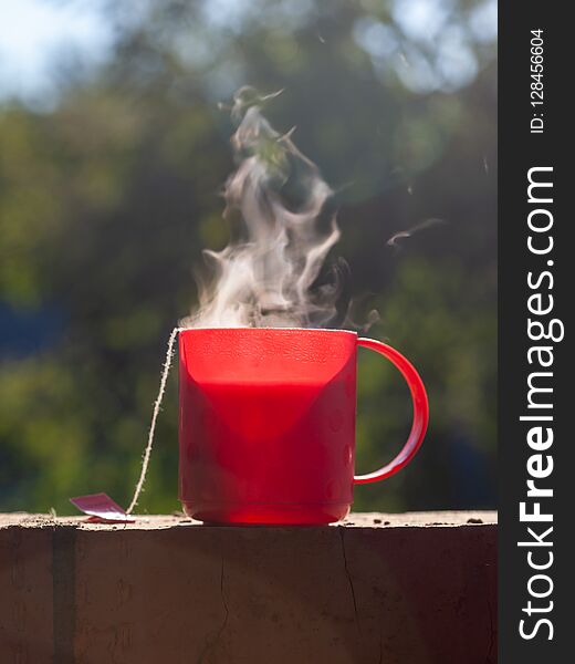 A steaming red mug of hot tea outdoors at natural background. A steaming red mug of hot tea outdoors at natural background