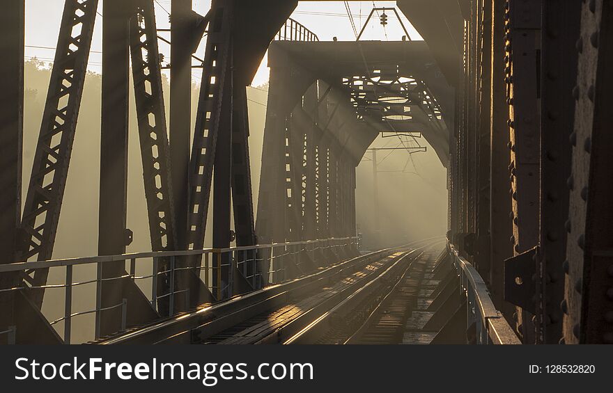 A Railway Bridge In The Morning Fog Or Smoke Through Which The Rays Of The Sun Shine