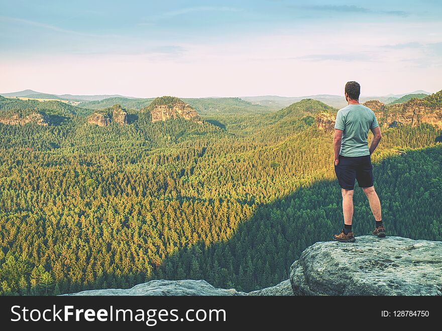 Tall man in outdoor clothes stands alone on the peak of rock. Hiker watching hilly forest landsape in morning mist.