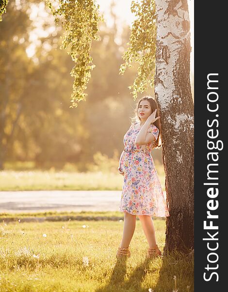 Pregnant girl in a light dress is walking around the park leaning against a tree. Concept maternity, pregnancy, childbirth, a healthy walk in the park, nature help. Sunset