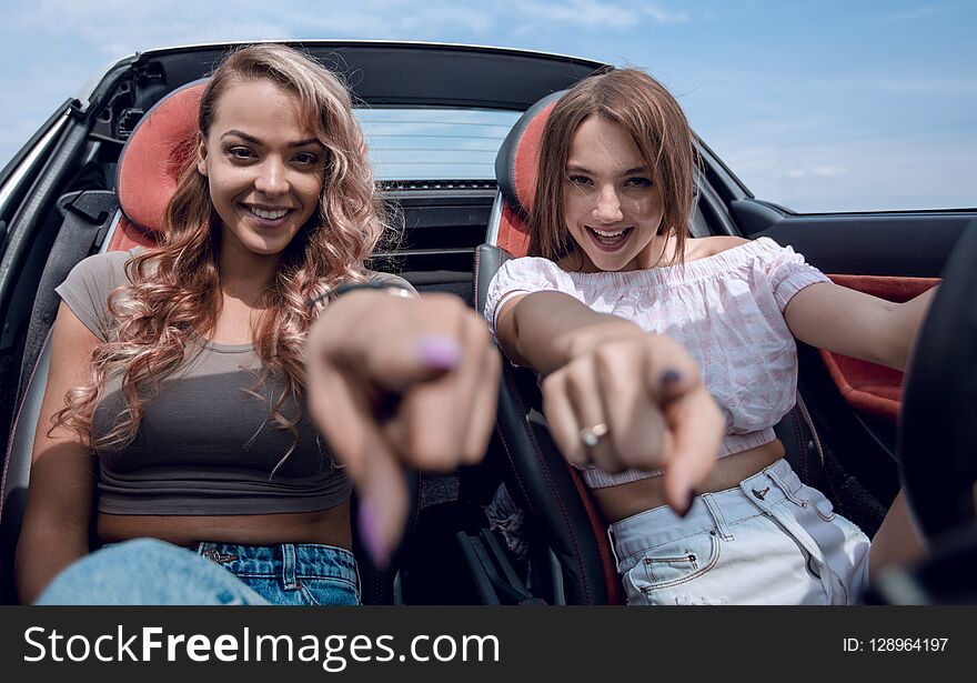 Two happy young women sitting in a car and pointing at you.