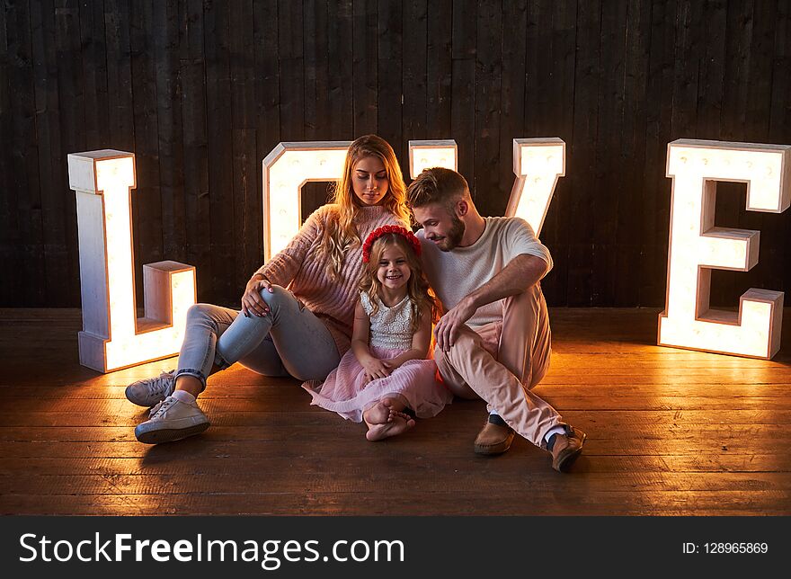 Mom, dad and daughter sits together in room decorated with voluminous letters with illumination.