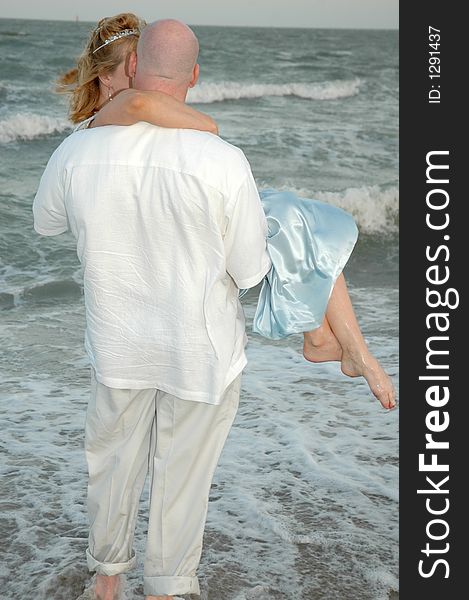 Happy groom picks up his bride. Man holding his woman in his arms on the beach shore. Standing in the waves of the ocean. Couple facing the ocean. Happy groom picks up his bride. Man holding his woman in his arms on the beach shore. Standing in the waves of the ocean. Couple facing the ocean.