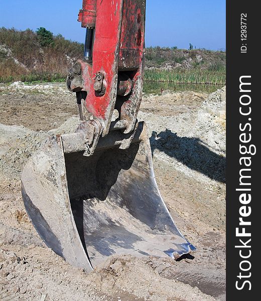 The image shows an excavator shovel of a red digger, digging in the sand. The picture was taken in a gravel-pit, blue sky is in the background.