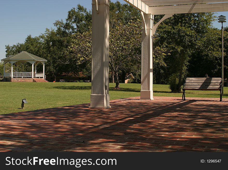 Picture taken from a covered patio in a small- town city park. Park benches, a gazebo and a birdhouse are in the background.