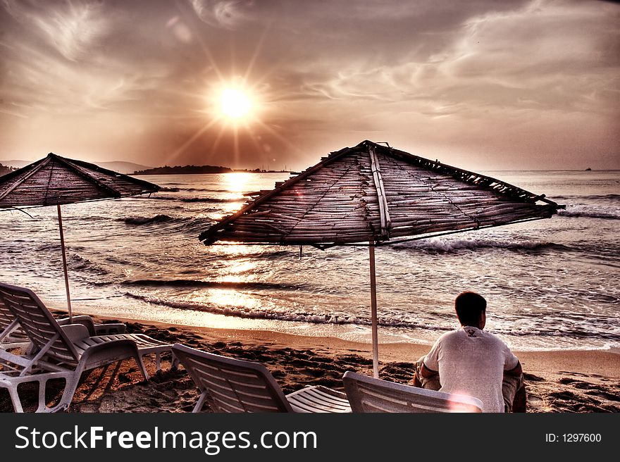 Man sitting in lounge under sunshade and looking at the sea at sunrise. Artistic photo. Noice visible at 100%. This is HDR photo with vivid and warm colors. Man sitting in lounge under sunshade and looking at the sea at sunrise. Artistic photo. Noice visible at 100%. This is HDR photo with vivid and warm colors.