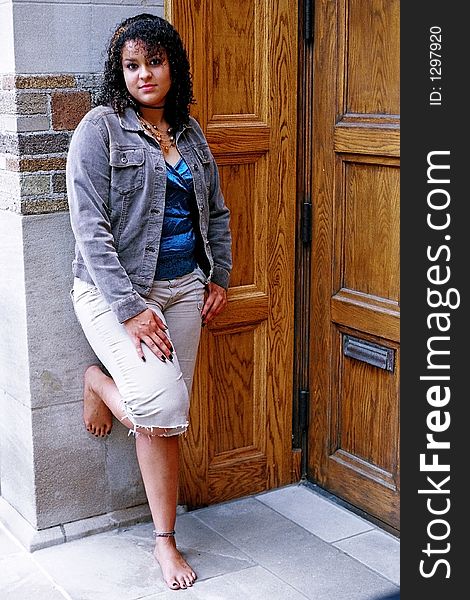 Woman posing in front of door waiting to go in, with one hand on thigh, one hand in pocket and right foot up on wall behind