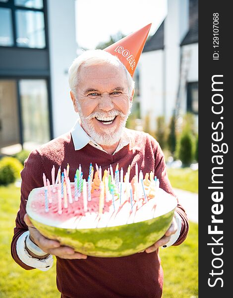 Funny celebration. Cheerful pensioner wearing paper hat while demonstrating his watermelon with candles. Funny celebration. Cheerful pensioner wearing paper hat while demonstrating his watermelon with candles