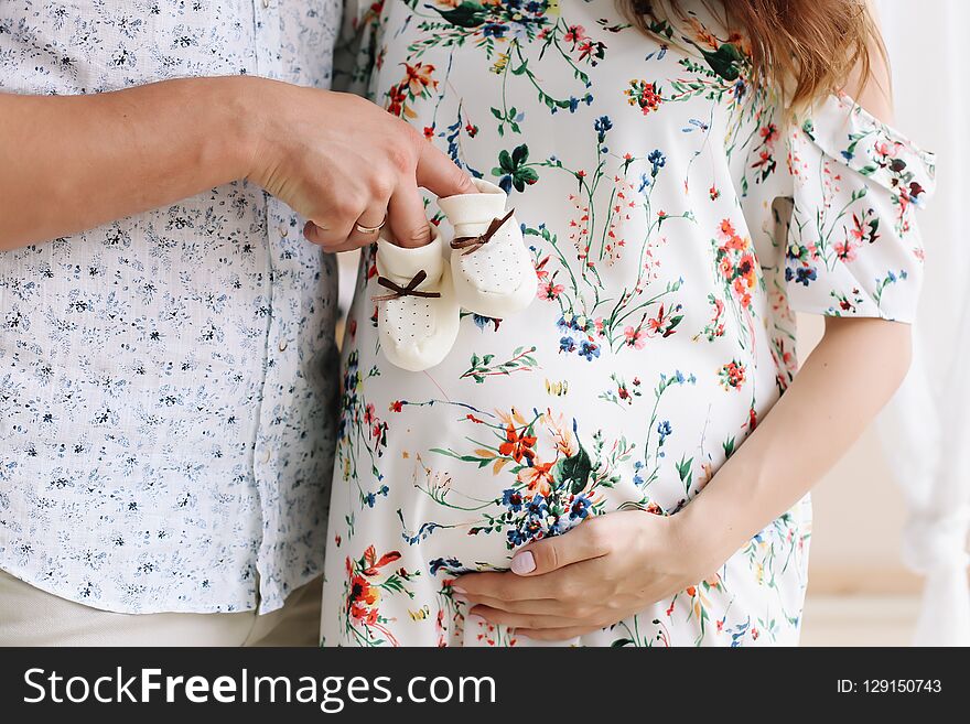 Conceptual photo of couples hands holding baby shoes. Pregnant woman in dress on white background. Pregnancy, maternity concept.