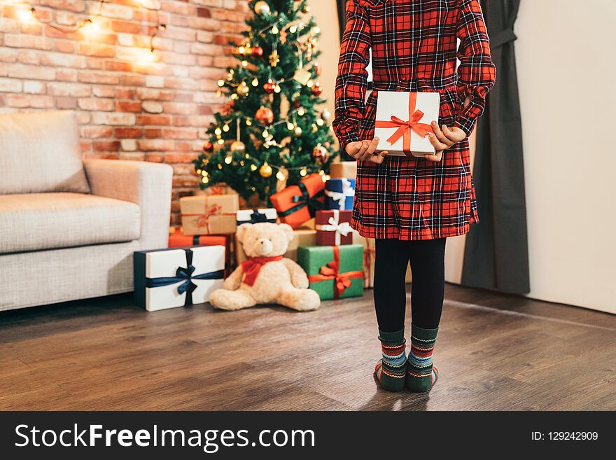 Little girl child holding gift in her back in the living room. attractive girl looking at the gifts and the teddy bear under christmas tree. cute kid with colorful socks standing on the wooden floor.