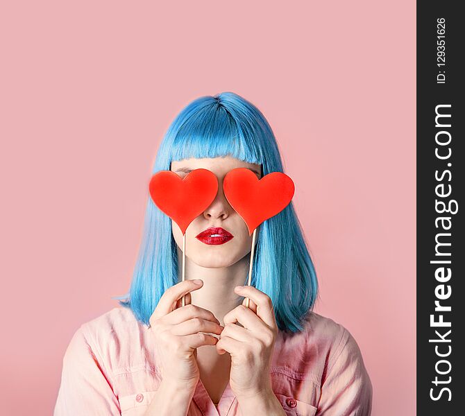 Young stylish woman with blue hairstyle and red lipstick holding valnetine`s heart on her eyes. Hipster funny girl model over pink pastel studio background. Love and valentine`s day design concept.