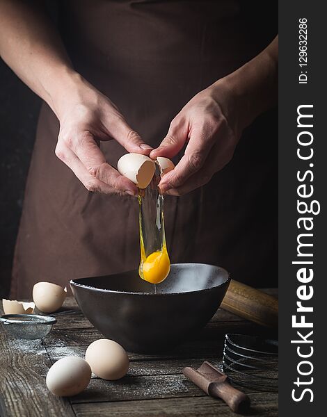 Professional chef hands are breaking an egg into bowl to make dough on wooden table, over dark background
