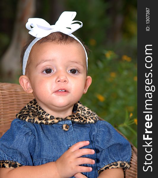 A cute little girl with a big bow in her hair sitting in a child's wicker chair wearing a denim outfit trimmed in leopard print. A cute little girl with a big bow in her hair sitting in a child's wicker chair wearing a denim outfit trimmed in leopard print