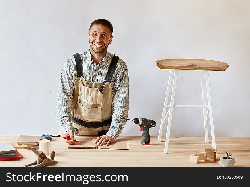 Carpenter smiling at camera while working with technical drawing lying on at studio, surrounded with carpentry tools and wooden planks, furniture making or house renovation process. Carpenter smiling at camera while working with technical drawing lying on at studio, surrounded with carpentry tools and wooden planks, furniture making or house renovation process