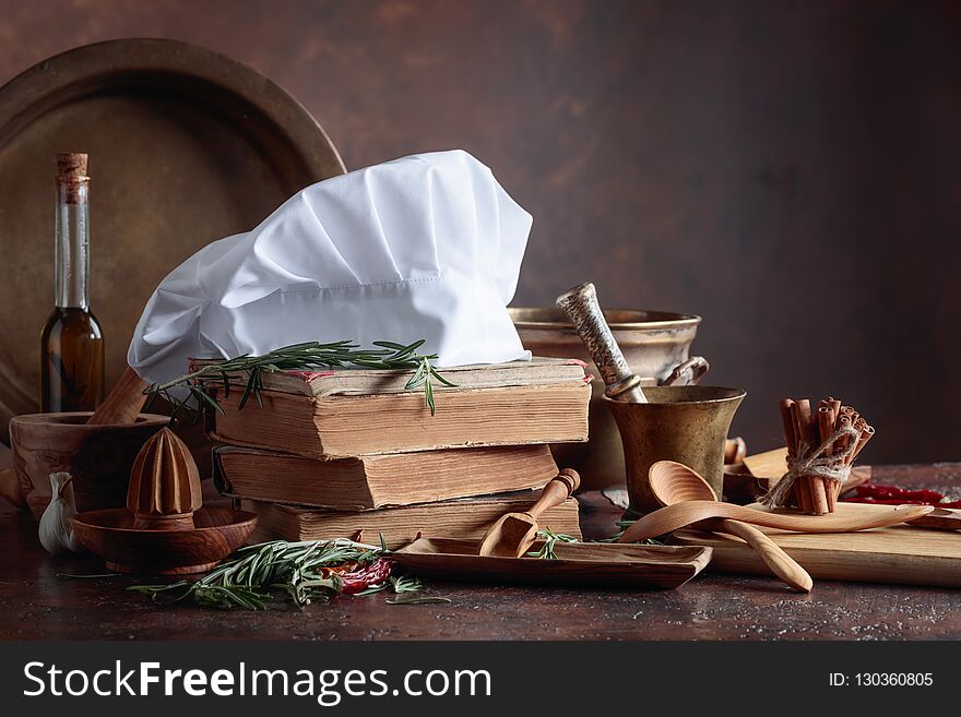 White chef`s hat and old cookbooks. Kitchen utensils with spices and rosemary on a kitchen table.