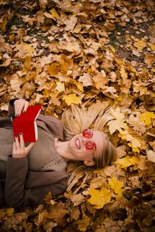 Beautiful Young Blonde In Pink Glasses Lies In Yellow Autumn Leaves, Reading A Book In Red Cover Royalty Free Stock Image