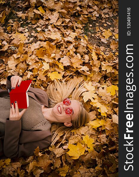Beautiful young blonde in pink glasses lies in yellow aun leaves, reading a book in red cover, copyspace, vertical photo. Beautiful young blonde in pink glasses lies in yellow aun leaves, reading a book in red cover, copyspace, vertical photo