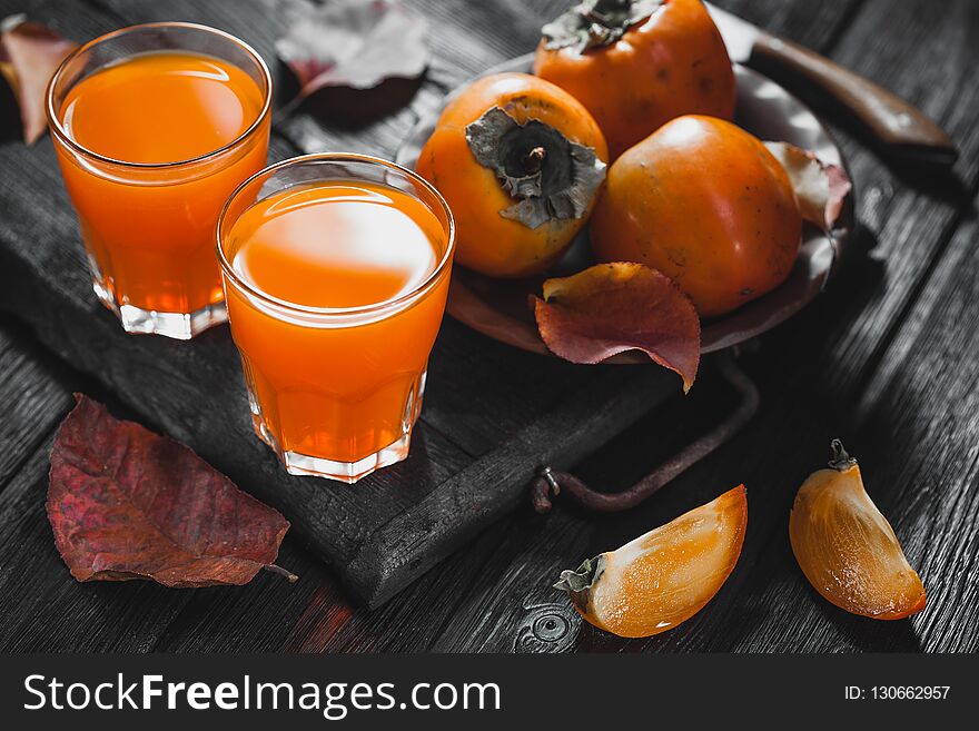A glass of fresh juice and ripe orange persimmon fruit and persimmon leaves in a brown plate on a black wooden table. fresh fruits