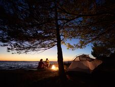 Young Couple Man And Woman Having Rest At Tourist Tent And Burning Campfire On Sea Shore Near Forest Stock Photo