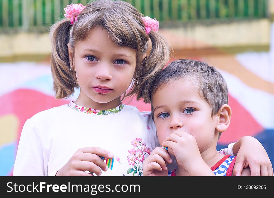 Little Caucasian Boy And A Girl Chewing Gum While Standing On The Playground Arm In Arm. Image Of Happy Friends Having