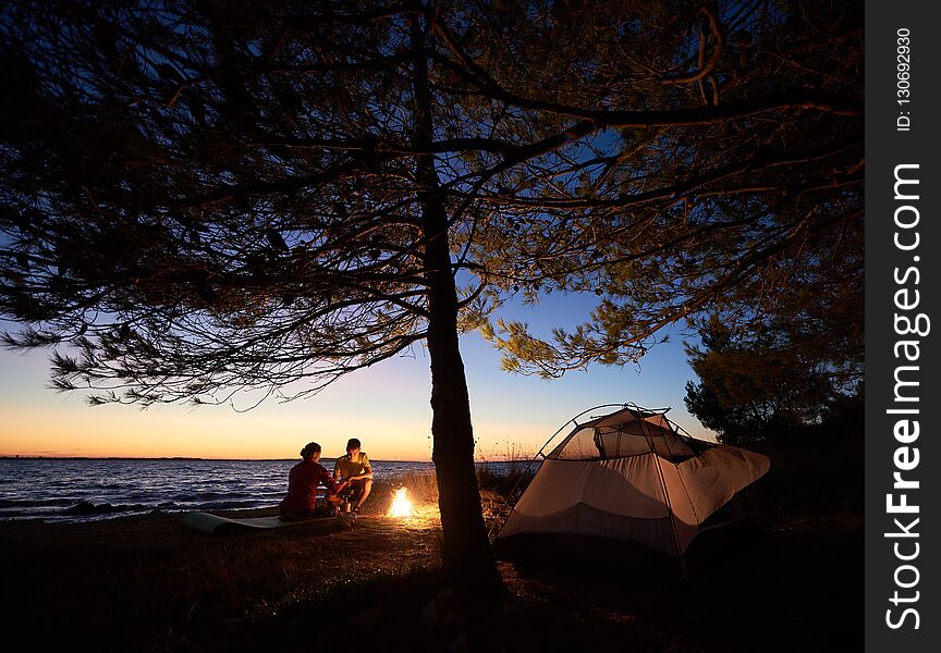 Young couple man and woman having rest at tourist tent and burning campfire on sea shore near forest