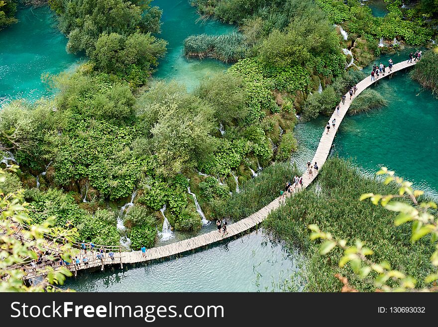 Plitvice Lakes National Park - UNESCO World Heritage Centre. Aerial view, colorful spring panorama of lit by sun green forest and long wooden bridge with tourists over blue lake with blue-green water.