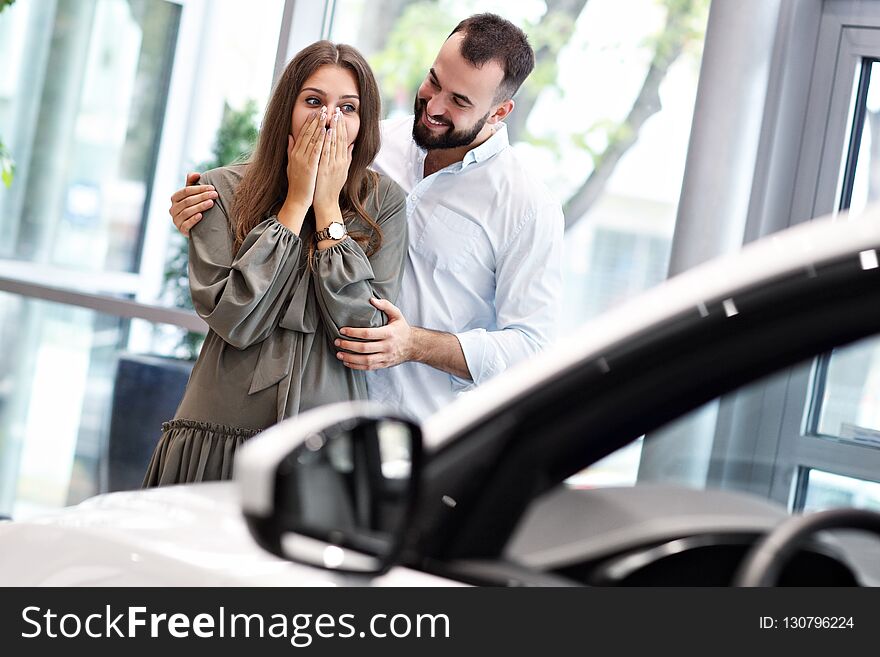 Adult man making surprise to beautiful woman in car showroom