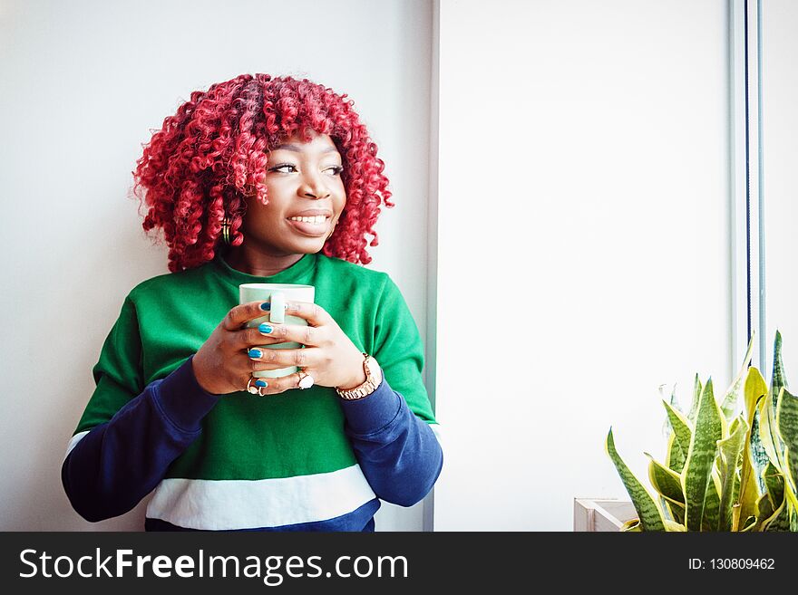 Young hipster dark-skinned woman dressed in casual clothes holding cup of hot drink, enjoying coffee or tea