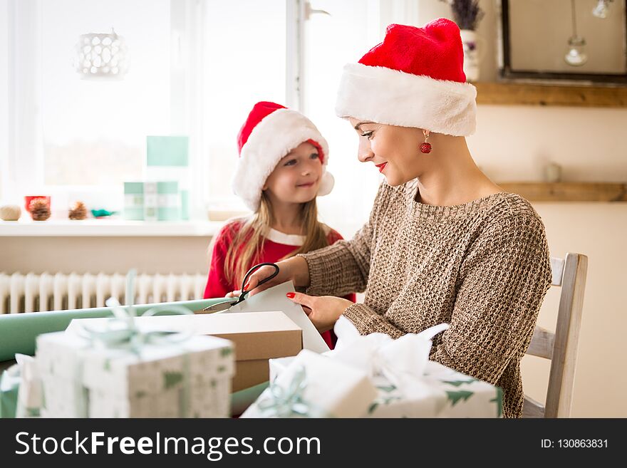 Mother and daughter wearing santa hats having fun wrapping christmas gifts together in living room. Candid family christmas time lifestyle background.