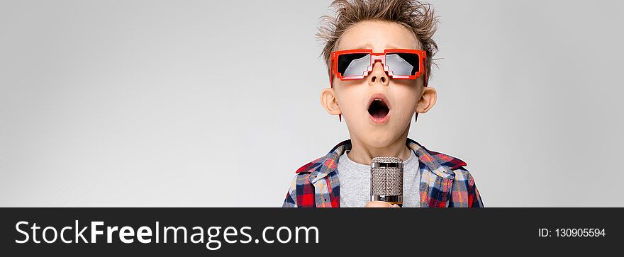 A handsome boy in a plaid shirt, gray shirt and jeans stands on a gray background. A boy wearing sunglasses. Red-haired