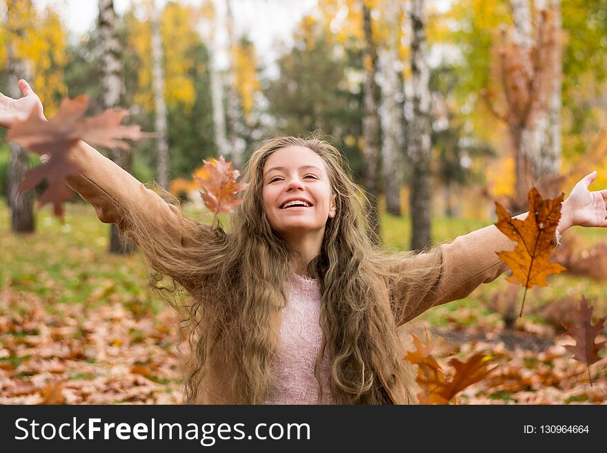 A teenager girl in a coat laughs and throws up leaves in autumn