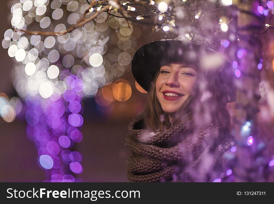Young girl standing in front of Christmas tree lights at night with snowflakes falling, snowing