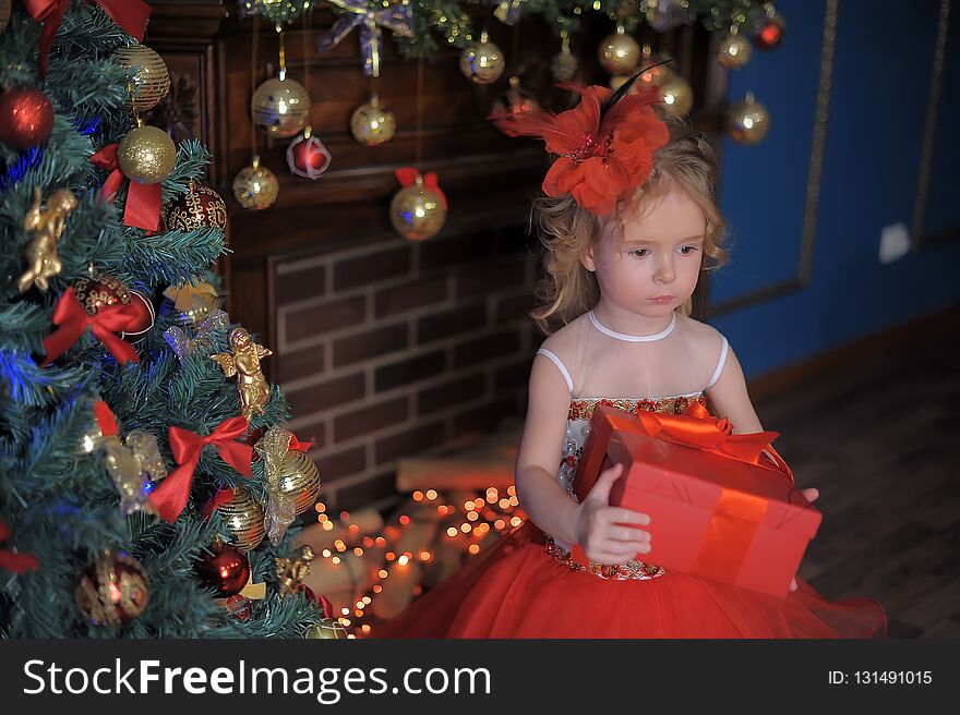 Cute little girl at the Christmas tree in a red elegant dress with a box with gifts in her hands