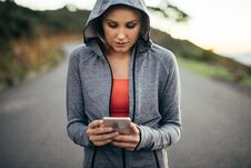 Fitness Woman Walking On Street Wearing A Hooded Sweat Shirt Looking At Her Mobile Phone. Woman Using Her Cell Phone Walking On An Stock Image