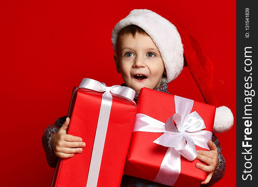 Funny surprised joyful child boy in Santa red hat holding Christmas gift in hand over the red background. Funny surprised joyful child boy in Santa red hat holding Christmas gift in hand over the red background